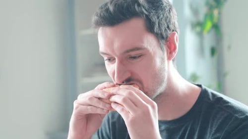 Young caucasian man bites into burger at home, cheeseburger eating enjoyment, close up portrait fast