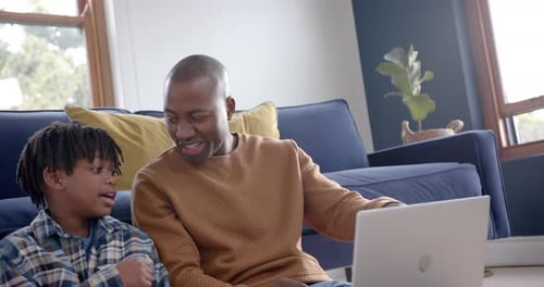 Happy african american father and son sitting on floor and using laptop at home, slow motion