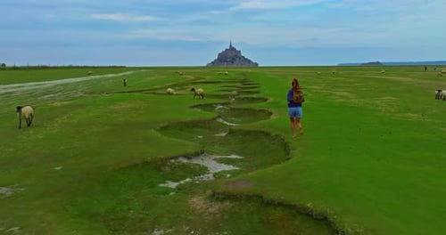 A Female with a Backpack Visits the Mont Saint Michel Castle While Walking Through Green Meadows