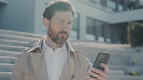 Serious Attractive Face Stylish Man with Beard in Coat Uses Smartphone Stand in Part Near Lake at