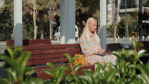 Young Beautiful Blonde Woman is Sitting on a Park Bench and Working on a Laptop
