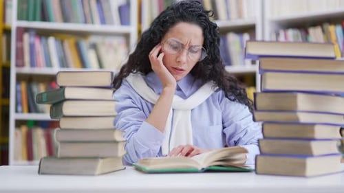 Tired female student studying by reading books in campus library space. Exhausted brunette girl