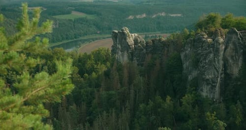 A towering rock formation stands amidst a dense forest, with a river winding through the lush valley