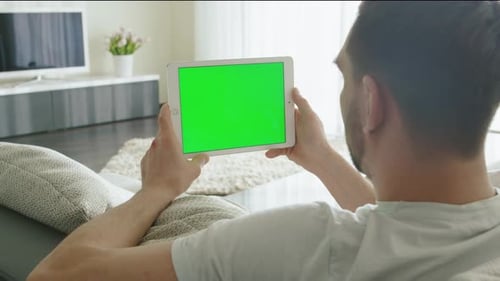 Man is Laying on Couch at Home and Watch on Tablet with Green Screen in Landscape Mode.