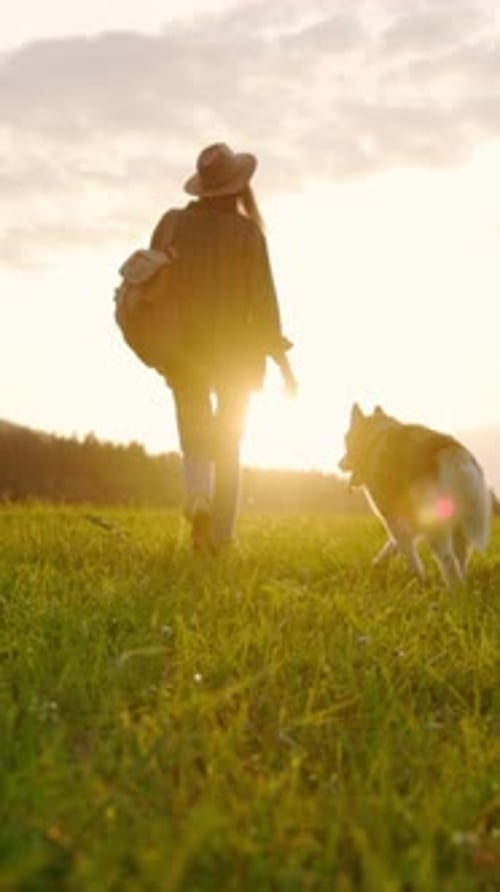 Woman and Dog Walk Through Grassy Field at Sunrise