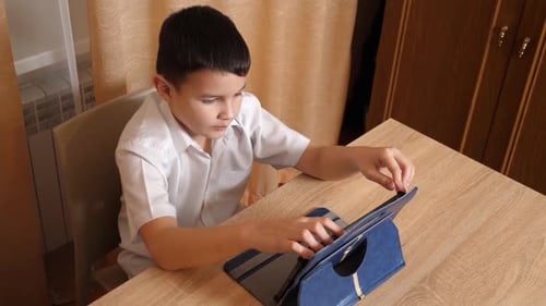 55. A boy plays games on a tablet while sitting at a table.