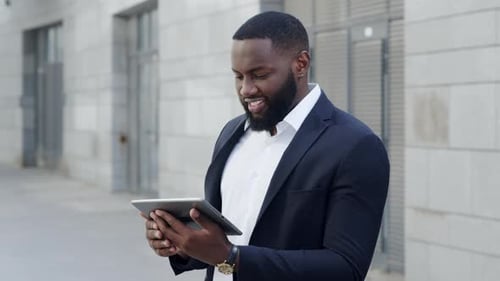 Smiling african businessman works on tablet at city street in slow motion