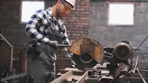 Male factory worker in hard hat and uniform is cutting metal pipes by a big saw