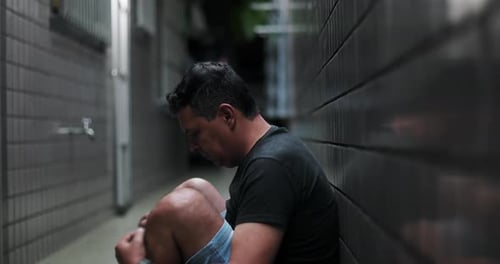Man sitting against a wall in a narrow alley, head lowered, struggling with inner turmoil,