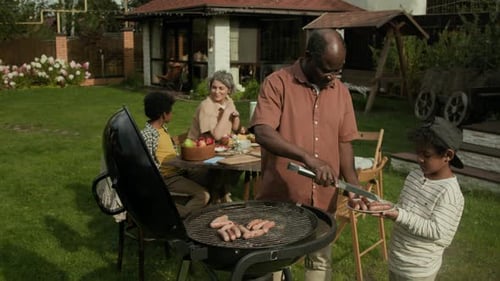 Black Father and Son Taking Hot Roasted Sausages off Barbecue at Garden Party