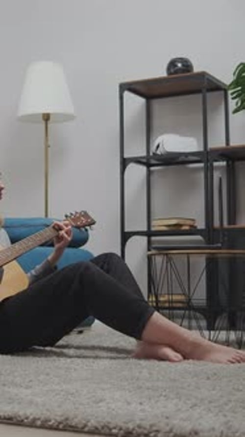 Teen Girl Sitting on Living Room Floor Playing Acoustic Guitar Near Blue Sofa and Window Practicing