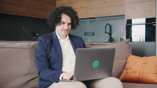 Curly Focused Business Man in a Jacket and Pajama Pants Working on Laptop Sitting at Home Office