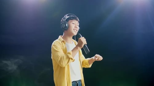 Side View Of Young Asian Boy With Headphone Holding A Microphone And Singing On Black Background