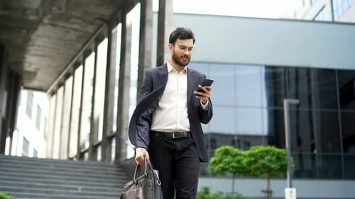 Smiling businessman walking on street holding smartphone outside office building. Confident handsome