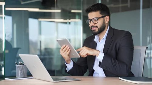 Smiling businessman is using browsing digital tablet sitting at workplace in business office.