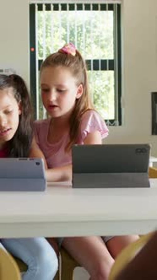 Vertical video: Reacting to cue, two girls in pink tapping blue tablet at school table, gray tablet