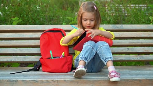 Little Girl Sits on Bench with Backpack in Schoolyard and Does Homework
