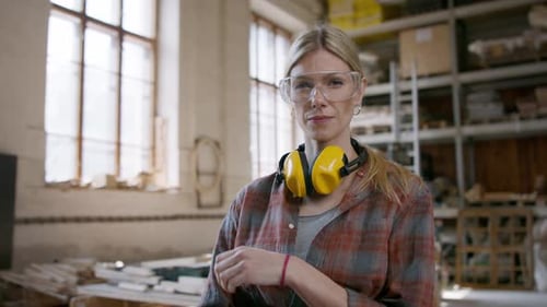 Female carpenter with goggles looks confidently at camera in her workshop