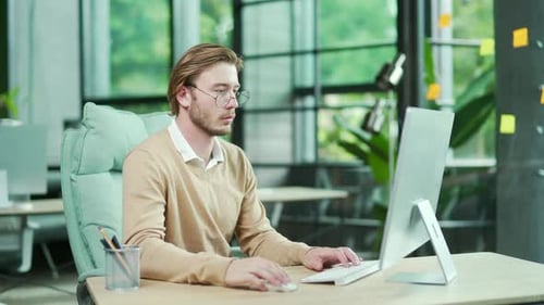 Jeune homme d'affaires concentré, employé avec des lunettes, travaillant sur ordinateur, vérifiant ses courriels dans un travail moderne