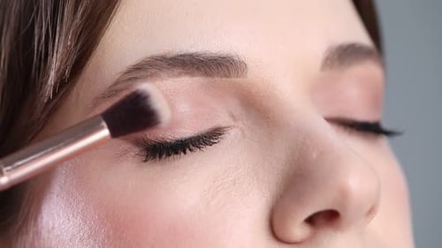 Applying nude eye shadow with makeup brush onto woman's face, closeup