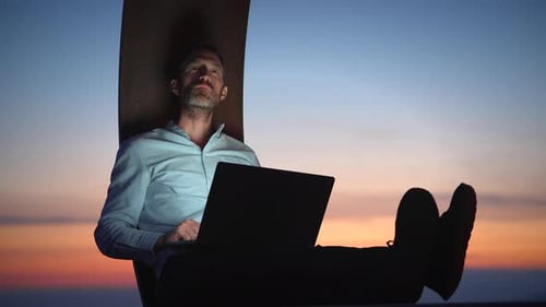 Ein ruhiger Arbeitsplatz am Abend Ein Mann, der mit seinem Laptop vor einem wunderschönen Sonnenuntergang tief nachdenkt