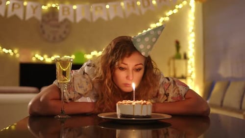 A Woman in a Nice Dress and a Party Cap Lays Hear Head on the Table and Blows Candles on the Cake