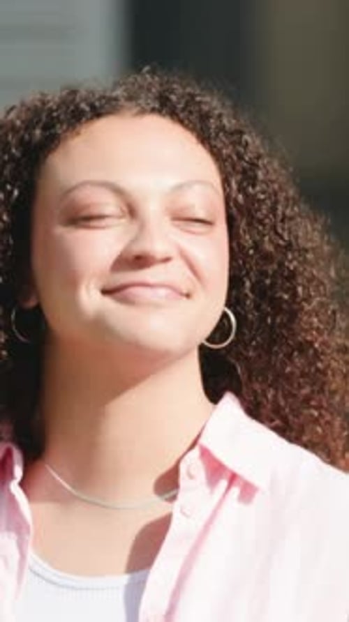 Vertical video: Turning curly-haired woman checking behind, walking away at sunny wall, pink shirt