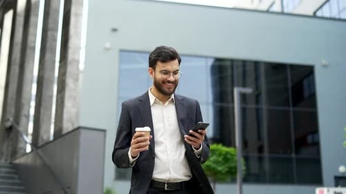 Smiling businessman walking on street holding smartphone and cup of coffee in hands outside office