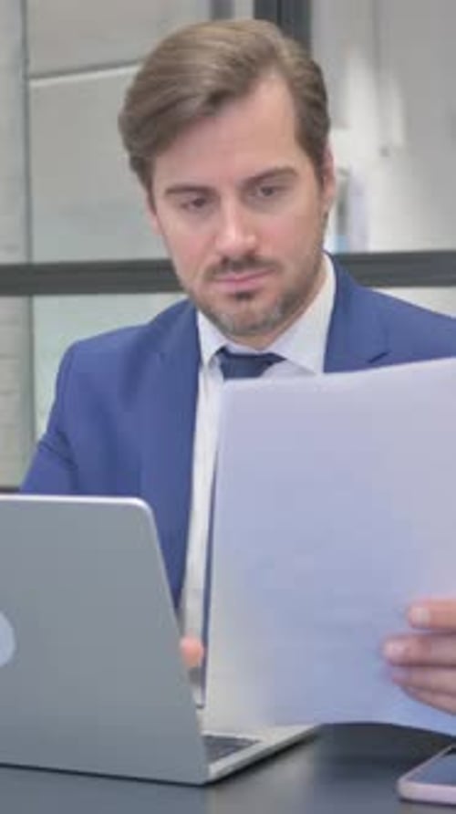 Professional Man Reviews Documents at Desk with Laptop