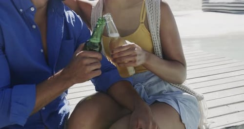 Happy diverse couple smiling and drinking a toast with beers on beach sun deck, in slow motion