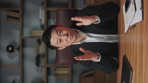 Asian Businessman Waving Hello on Camera at Office Desk