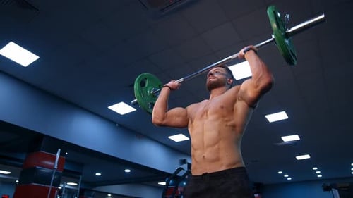 Ein gutaussehender männlicher Bodybuilder hebt die Barbe über den Kopf. Selektiver Fokus auf Sportler ohne Hemd. Zeitlupe.