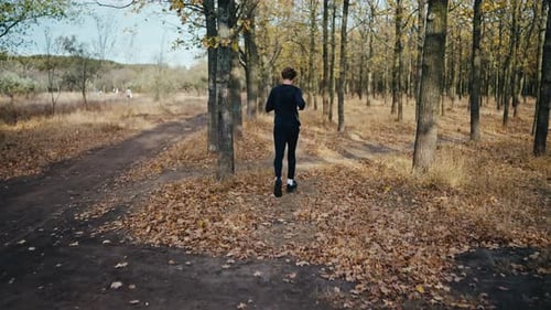 A Brunette Man with Curly Hair in a Black Sports Uniform Runs Through the Autumn Forest Along an