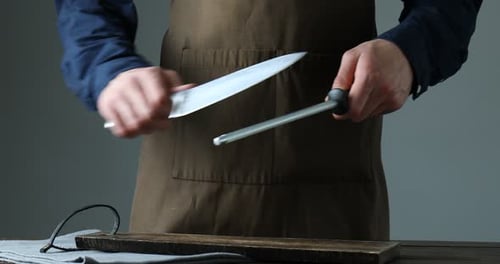 Man sharpening knife with sharpener at wooden table against grey background, closeup