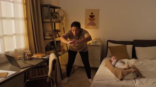 Young Mother Exercising at Home to Fitness Video and Baby Lying in Cocoon