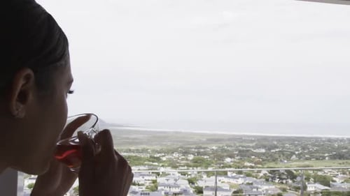 Thoughtful biracial woman looking through window drinking tea at home, copy space, slow motion