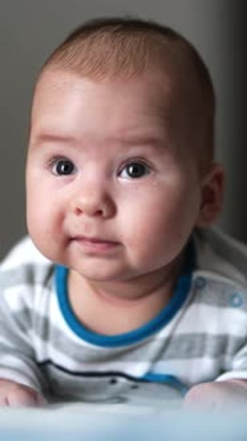Beautiful little toddler lies on the bed on his belly. Sweet kid grimacing funnily and looking up.