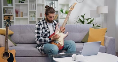 Talented musician conducts class on laptop sitting on sofa in modern studio playing guitar. Caucasia