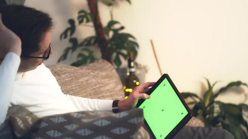 Close up of man lying down on sofa watching digital tablet with chroma key during winter eve. Green