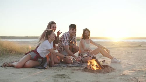 Amigos curtindo uma fogueira na praia ao pôr do sol assando marshmallows e compartilhando histórias em um ambiente aconchegante e