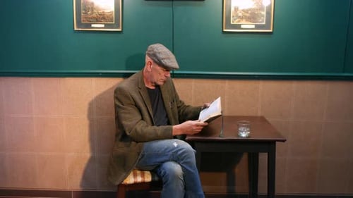 A lonely man wearing a flat cap is seated at a pub table, intently reading a book. A glass sits on t