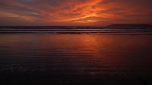 Seascape view of ocean waves crashing on the sandy coastline, during a bright red and orange sunset,