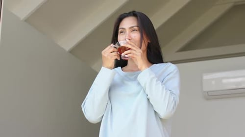 Holding glass tea mug, Asian woman sipping slowly and finding calm beneath slatted balcony roof