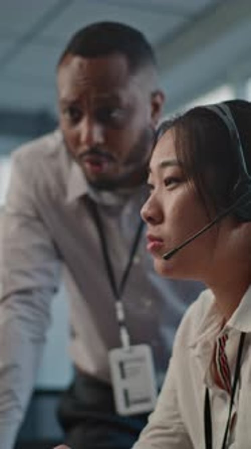 Asian Helpdesk Specialist in Headset Uses Computer Talks to African American Colleague