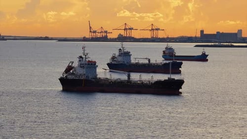 Aerial drone view of multiple oil tankers anchored near an industrial port at sunset with cranes and