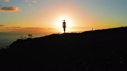Silhouette einer einsamen Frau auf einem Berggipfel während eines Drohnenflugs bei orangefarbenem Sonnenuntergang