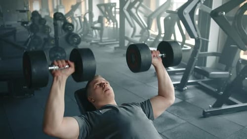 Mittelgroße Aufnahme eines jungen Sportlers beim Bankdrücken mit Hanteln in einem ausgestatteten Fitnessstudio