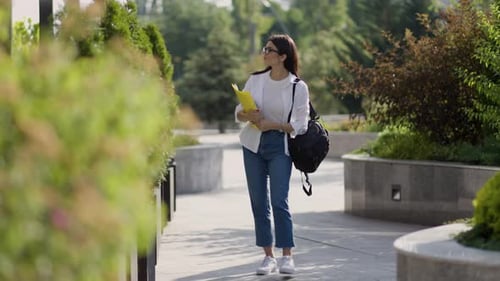Happy Young Woman Standing with Books and Backpack on Street in Sunlight