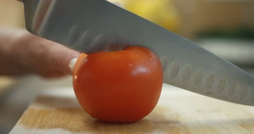 Close-up Shot of a Person Cutting Tomato in Half with Sharp Knife on a Wooden Board.