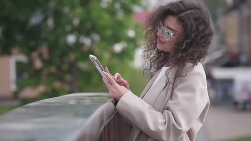 A Young Woman with Curly Hair Stands on the Street in the Summer and Writes a Message on a Gadget